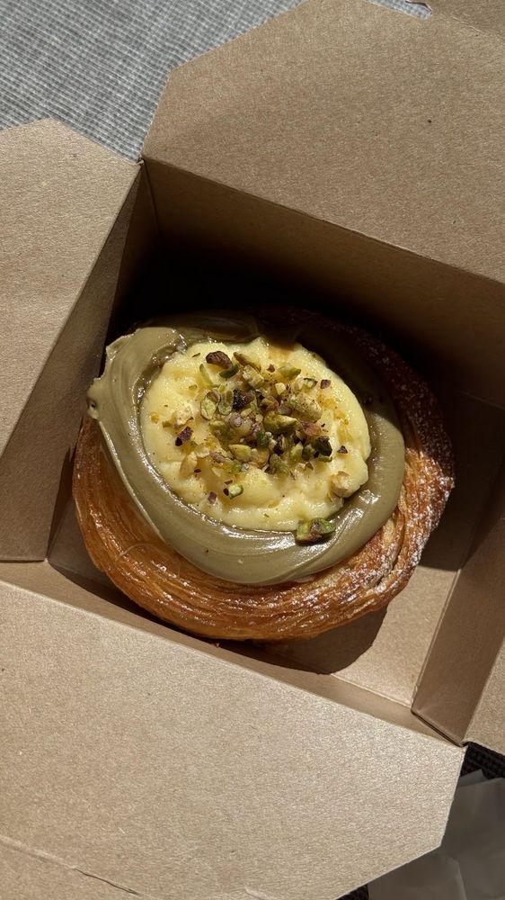 A pistachio-cream filled round croissant in an open kraft takeaway box